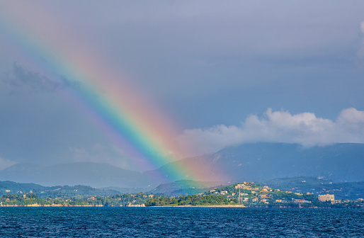 Beautiful summer landscape with sea, small village on the hill, bright colorful rainbow on blue sky, clouds and mountains on the horizon. Corfu Island, Greece