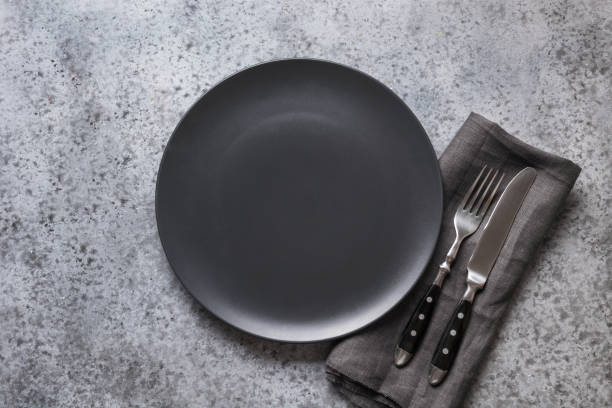 istock Black plate, fork and knife on grey stone table. Minimal table place setting. Space for text or menu. View from above. 1203208075