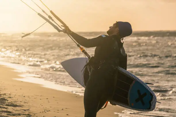 Photo of Portrait of a wave kitesurfer walking upwind at the beach with his board and a kite in beautiful yellow sunset conditions