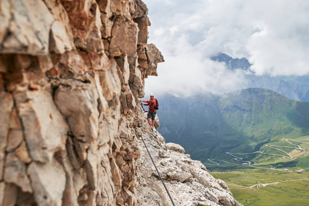 homme sur la via ferrata cesare piazzetta, montagnes de dolomites, italie, près du mur de roche, avec une route sinueuse ci-dessous, un jour d’été avec des nuages se rassemblant rapidement derrière. - european alps mountain mountain peak rock photos et images de collection