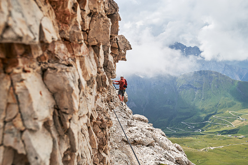 Man on via ferrata Cesare Piazzetta, Dolomites mountains, Italy, close to the rock wall, with a winding road below, on a Summer day with clouds gathering fast behind.