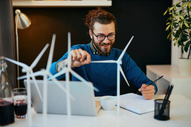 istock Young creative caucasian bearded environmentalist  with curly hair sitting in his office, touching windmill model and drawing new better model in notebook. 1203219164