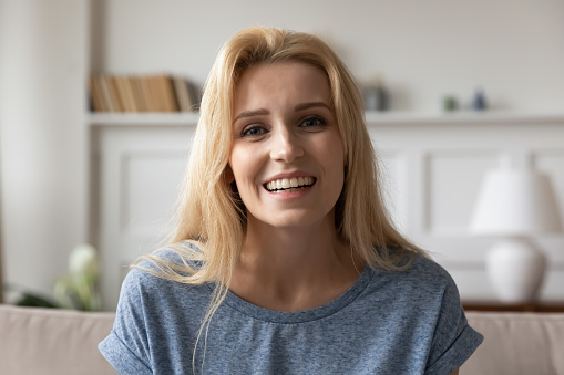 Head shot close up portrait of attractive smiling young blonde woman. Pleasant millennial girl looking at camera, holding video call with parents or recording vlog for personal channel at home.