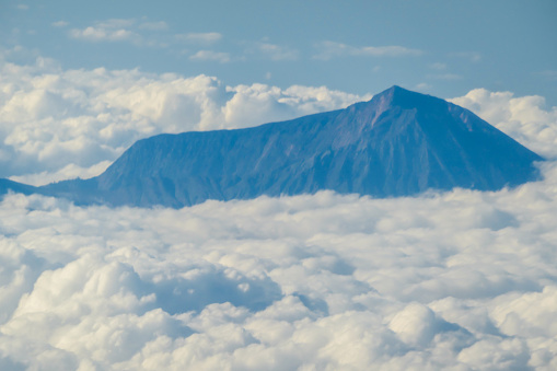 An island sneak peaking through the clouds seen from a plane's window, spotted while flying over Indonesia. Everything is covered with clouds except the mountain peak. Top down perspective.