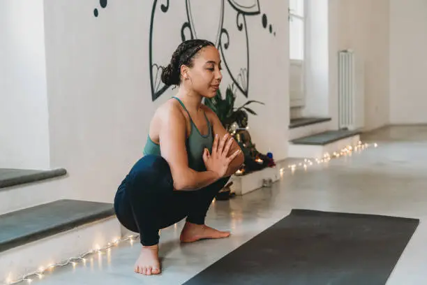 Young adult woman practicing yoga in garland position. She's meditating.