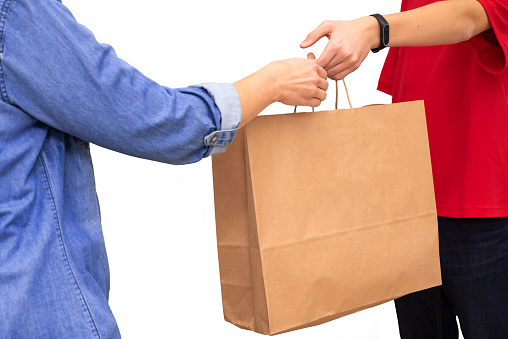 Female hand receiving package from delivery man. Young delivery courier dressed in red t-shirt holding brown paper bag isolated on white background. Delivery service concept. Copy space