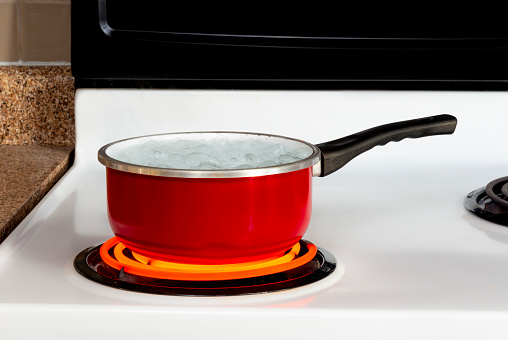Horizontal shot of a red pan of boiling water on top of a stove with the burner turned to high.