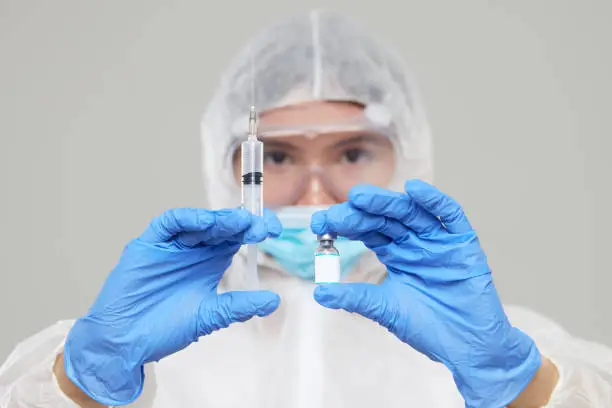 Photo of Biological hazard. Epidemic of the Chinese coronavirus. An asian woman in a protective suit and mask holds an injection syringe and vaccine. Vaccine from, flu, coronavirus, ebola, TB.