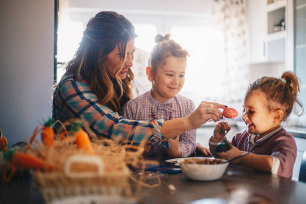 A mom and her two girls preparing painted eggs for Easter. Mom is teasing the younger one with a freshly painted egg. The daughter is laughing.