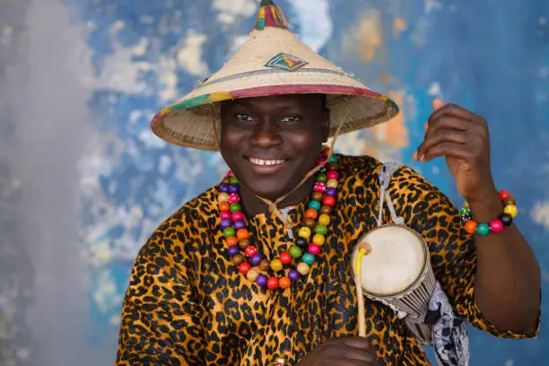 Photo of African man in traditional clothes and fulani hat playing on talking drum