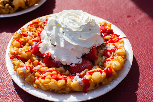 A funnel cake with strawberry syrup drizzle, whipped cream and powdered sugar sits on display.  This is food that is often popular at farmer's markets, carnivals, and fairs, and can also be made at home fairly easily.  It is a popular dessert, this serving size serves two to three people.