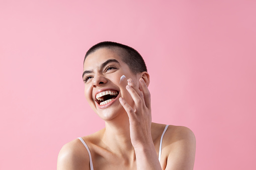 Excited young lady applying face cream on her cheek and laughing