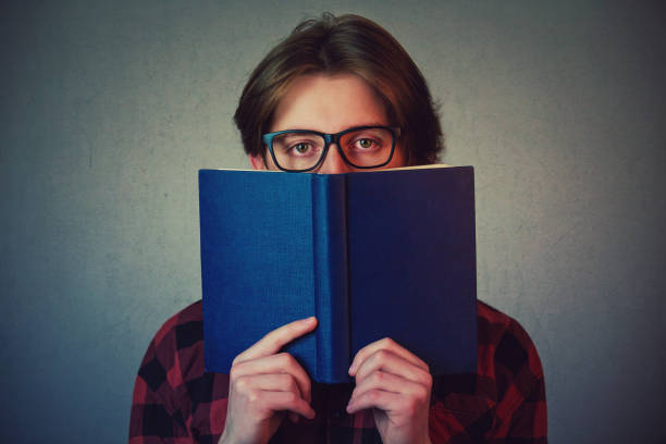 istock Close up portrait student boy hiding behind a opened blue book isolated on grey wall background. Mysterious, shy or scared teenage guy, nerd, bullied at school try to be invisible or incognito. 1203343147