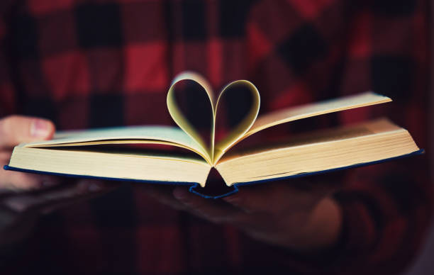 ページで作られたハートシンボルの形で開いた本を持つ読者の手をクローズアップ。学生の男の子としての愛の教育の概念は、灰色の壁の背景に隔離された教科書を読みます。 - hands open ストックフォトと画像