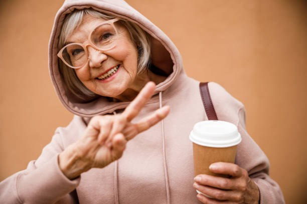 Happy elderly woman showing V sign stock photo Jolly aged female in hood and glasses is demonstrating peace gesture while drinking coffee grandma portrait stock pictures, royalty-free photos & images