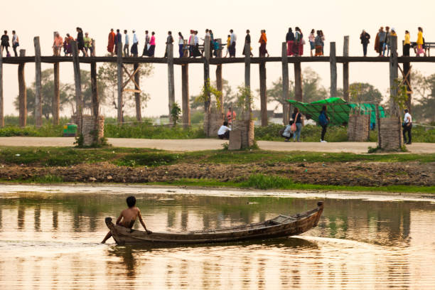 People Walking On U Bein Bridge And Watching Their Poor Boy On The Boat U Bein Bridge, Mandalaj, Myanmar - January 2018, A Poor Boy Sits On a Boat And Looks At People Walking On U Bein Bridge u bein bridge stock pictures, royalty-free photos & images