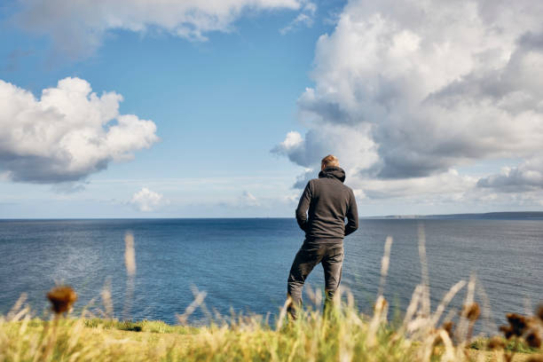 istock Man stood alone looking out over an idyllic bright sunny seascape at Pentire, Newquay, Cornwall on a Septembers day. 1203364113