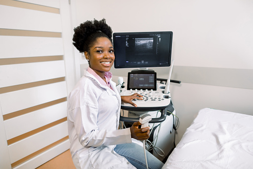 Doctora de mujer africana sonriente con escáner de ultrasonido en la mano, trabajando en la moderna máquina de escaneo de ultrasonidos en la sala de luz en la clínica. Retrato del operador de la máquina de exploración por ultrasonido 4D photo