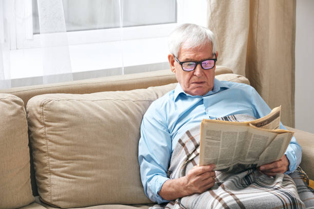 istock Senior man wrapped in plaid reading newspaper while relaxing on couch 1203365611