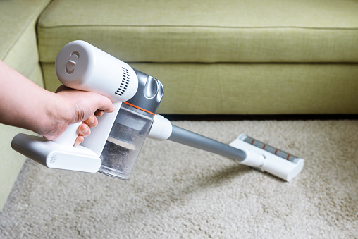 Wireless vacuum cleaner used on carpet in room. Housework with new upright hoover. Person holds modern white vacuum cleaner by sofa. Home cleaning, care and technology concept.