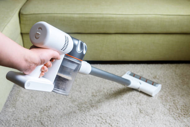 Wireless vacuum cleaner used on carpet in room. Housework with new white hoover. Person holds modern vacuum cleaner by sofa. stock photo