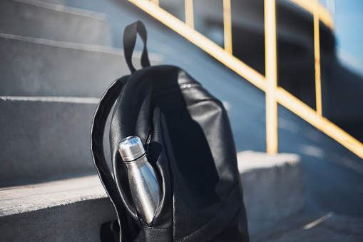 Close-up of black backpack with reusable steel thermo water bottle inside on urban stairs.