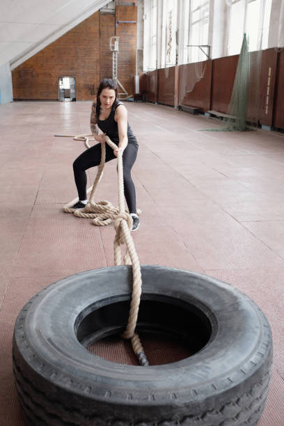 jeune athlète féminin caucasien tirant le pneu lourd avec la corde de bataille tout en ayant l'entraînement de formation de croix dans la salle de sports - fitness trainer photos et images de collection