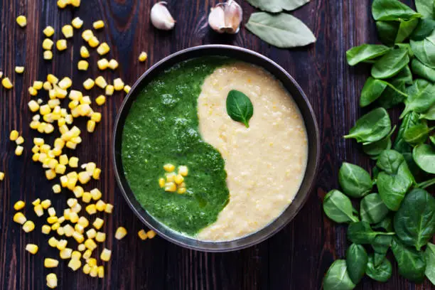 Assortment of spinach and corn cream soups on dark wooden surface, mixed as yin yang symbol