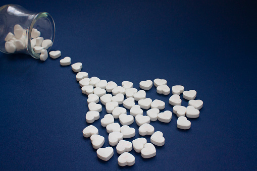 Many of white candy heart-shaped pills spilled out of a transparent jar on a dark blue background. in heart shape.