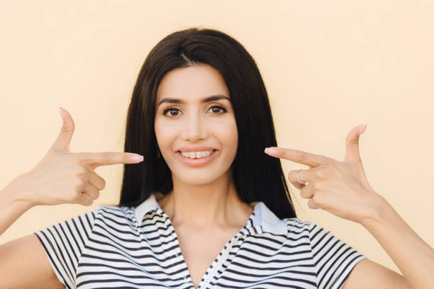 istock Portrait of pleasant looking brunette female has appealing look, indicates with both fore fingers at mouth shows gentle smile, teeth with braces, dressed in casual clothes, isolated on light wall 1203401288