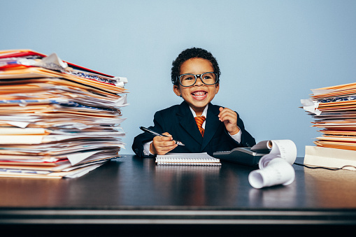 A young boy dressed in business suit and necktie, is sitting at his office desk playing tax accountant and financial advisor. On his desk sits many financial documents and a calculator. This boy is ready for crunch your numbers and make the most of your money.