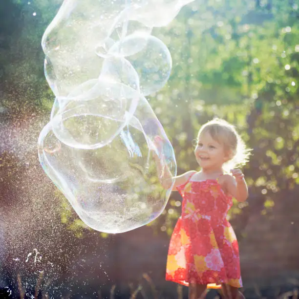 The cute little girl in dress is playing with giant soap bubbles outdoors.
