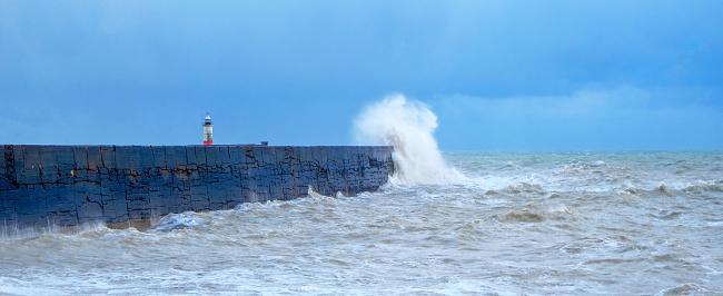 A harbor wall with a rough stormy sea crashing against the wall causing the sea to be blurred and in motion, behind is a red and white lighthouse, waves are crashing over the wall, there is a blue sky behind