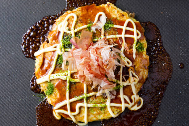 receta japonesa de okonomiyaki. espolvorea salsa, mayonesa, algas y bonito sobre okonomiyaki a la parrilla en un plato caliente. - japanese cuisine soy sauce food bonito fotografías e imágenes de stock