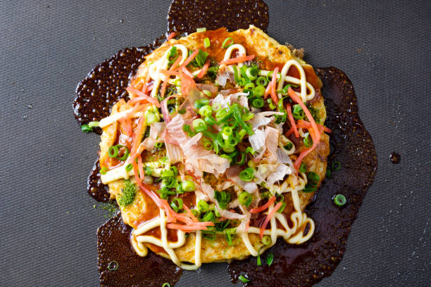 receta japonesa de okonomiyaki. espolvorea salsa, mayonesa, algas y bonito sobre okonomiyaki a la parrilla en un plato caliente. agregue las cebollas verdes y agregue el jengibre. - japanese cuisine soy sauce food bonito fotografías e imágenes de stock