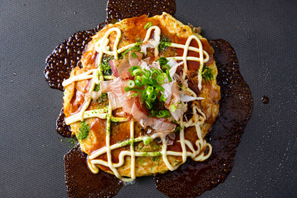receta japonesa de okonomiyaki. espolvorea salsa, mayonesa, algas y bonito sobre okonomiyaki a la parrilla en un plato caliente. agregue más cebollas verdes. - japanese cuisine soy sauce food bonito fotografías e imágenes de stock