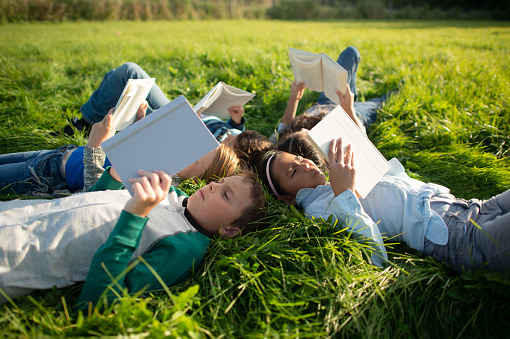 A group of multi-ethnic youth, lay outside in the grass, in a circle.  They are each dressed casually in long sleeve fall shirts and denim pants.  Each student is holding a book open overhead as they read together in the grass.