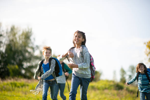 A group of multi-ethnic youth, run through a field on a warm sunny day.  They are dressed in layered fall clothing and have smiles on their faces as they run and have fun.  Each one has a backpack over their shoulders.