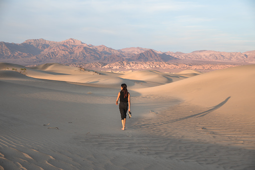Beautiful fit mixed race woman walking between large dunes in the desert of death valley national park, California. Holding sandals in hand, looking to side and long shadow cast to the right. Sunset.