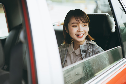Young Asian woman sitting in the backseat of a taxi looking out the window and smiling while commuting in the city