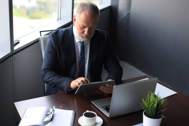 istock Mature business man in full suit using digital tablet while sitting in the office. 1203486653