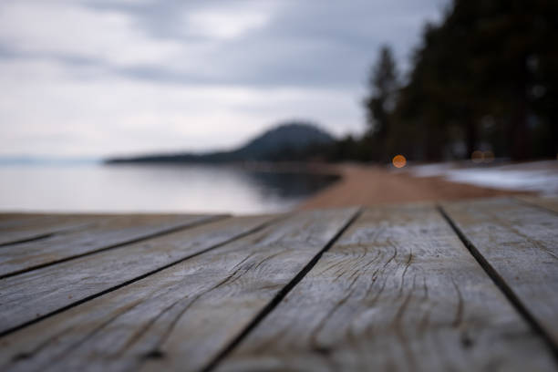 Close up of the weathered wood grain of a pier. Out of focus on the background is a peaceful evening scene with a lakeshore, pine trees and a mountain. Lots of copy space.