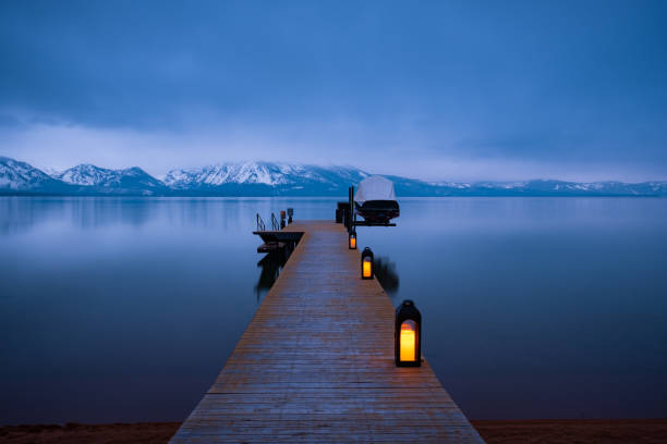 A weathered wooden pier extends out into the still blue evening waters of Lake Tahoe. Snow capped mountains and a stormy over cast sky reflect on the water.
