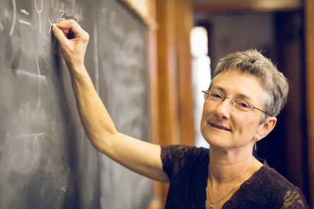 Senior female math teacher writing a formula to the chalkboard, looking over with a friendly smile. Personal Perspective. Natural interior light. Lunenburg, Nova Scotia, Canada.