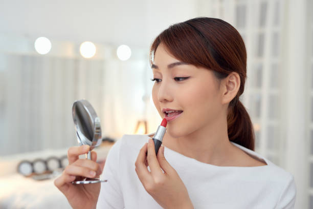 istock Smiling young Asian woman applying lipstick and looking to mirror. 1203520005