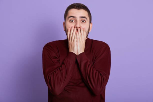 istock Portrait of scared impressed man with beard standing isolated over lilac background in studio, covering half of his face with hands, opening his eyes widely, being shocked. Emotions concept. 1203520303