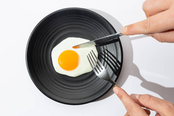 istock Hands are going to eat with knife and fork a fried egg on black plate on  white background. 1203525736