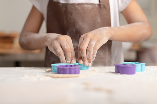 A little girl wearing a brown apron is using a blue mold to cut the dough for making cookies in the kitchen.