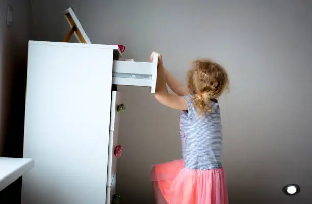 Photo of Young girl child climbing on modern high dresser furniture, danger of dresser dipping over concept. Children home hazards. Staged photo.