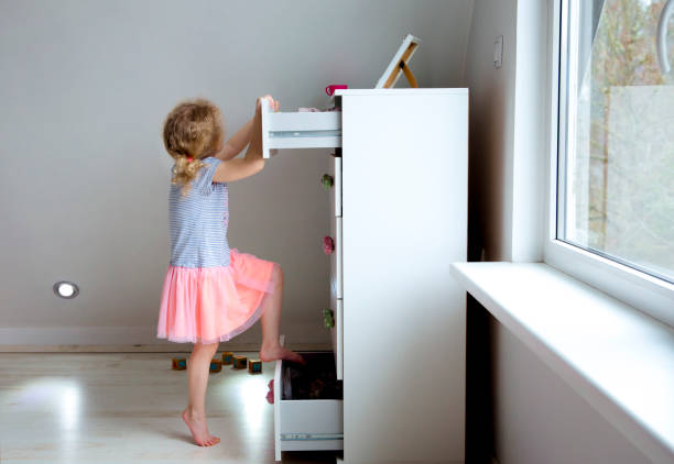 Young girl child climbing on modern high dresser furniture, danger of dresser dipping over concept. Children home hazards. Staged photo. Young girl child climbing on modern high dresser furniture, danger of dresser dipping over concept. Children home hazards. Staged photo. dresser stock pictures, royalty-free photos & images